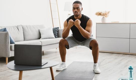 Person watching a fitness video on their laptop at home, doing a squat