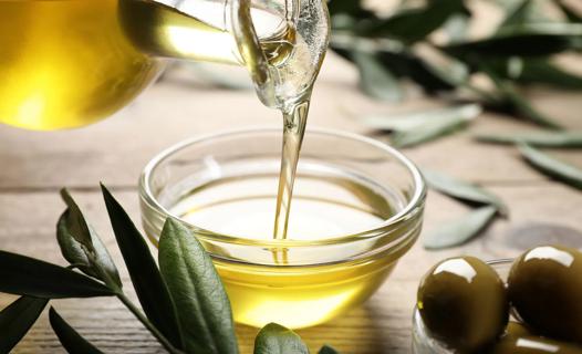 Olive oil being poured into small glass bowl from glass oil dispenser, with whole olives on table