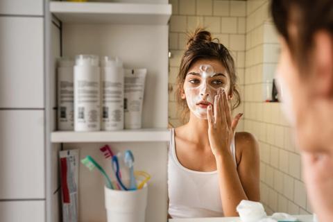 Person washing their face, using bathroom mirror, with shelves filled with skin care products